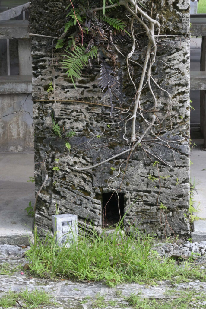 A photo of a small, collagraph sculpture posed at the base of a large column built of coral rock that has a rectangular alcove near the base which echoes the shape of the sculpture.