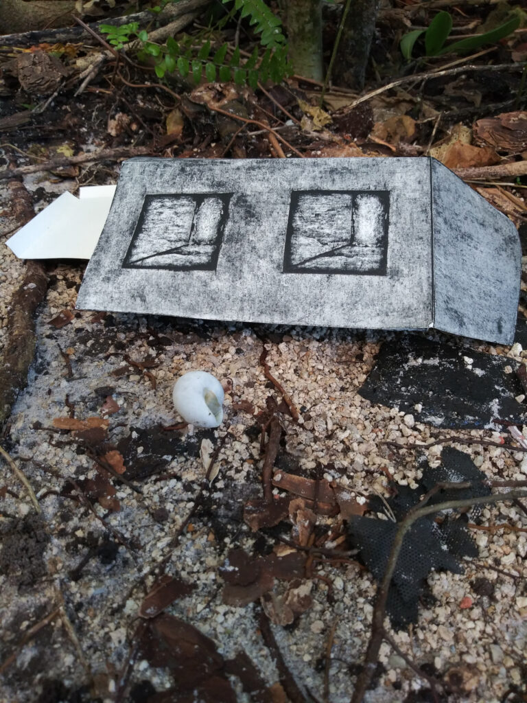 A photo of a flattened version of the collagraph 'cinder block' sculpture, laying among sand, detritus, leaves and a snail shell.