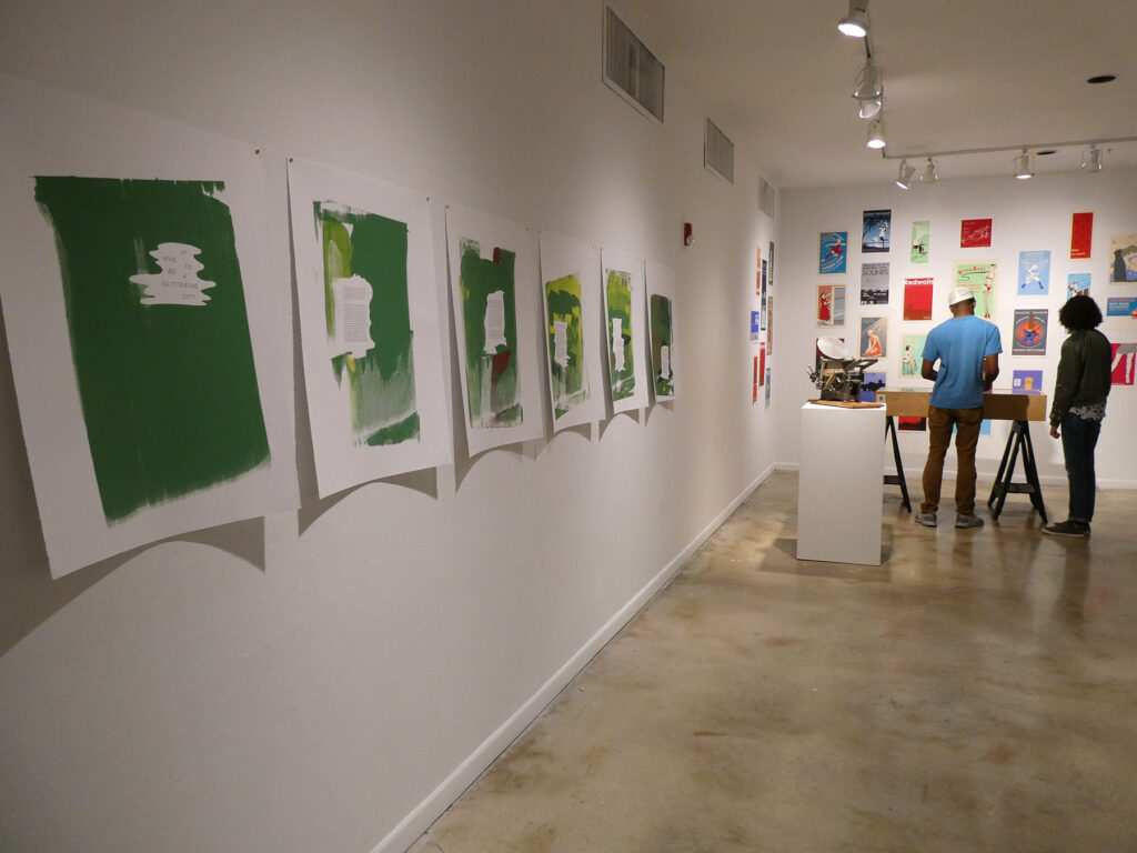 Image depicts a long wall with six large prints, a further wall with an arrangement of screenprinted posters, and two guests looking at the works.