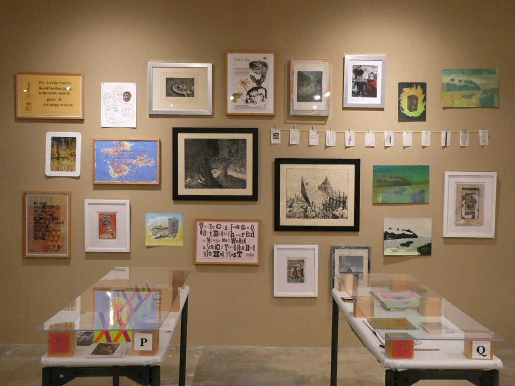 Image depicts a wall of prints arranged salon-style against a tan wall, with partial views of two vitrine tables in front.
