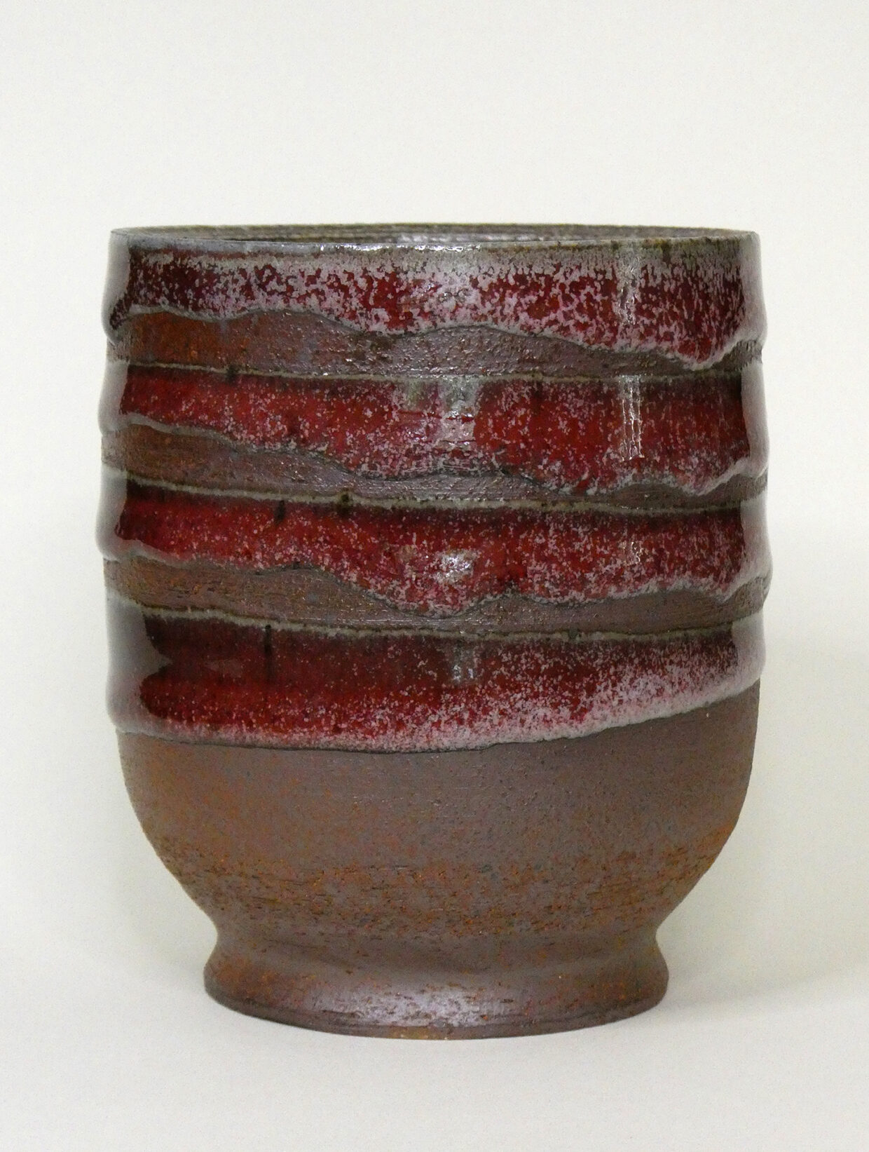 Photo of a small, soda-fired vessel with a rounded base, angled foot, no handle and red, drippy horizontal strips in four rows from the top, alternating with a dark clay body.